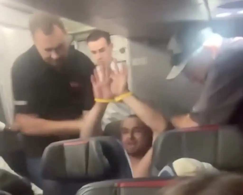 In the picture, the accused is seen sitting on the seat without a shirt. The flight crew members have tied his hands.