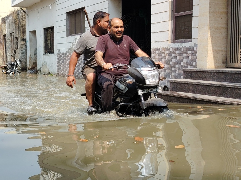 गली में भरे गंदे पानी के बीच से गुजरते बाइक सवार।