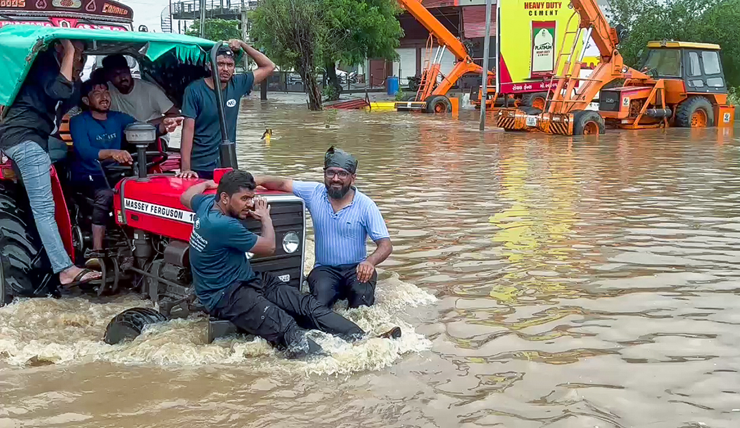 अहमदाबाद के बनासकांठा में रविवार को लगातार बारिश के बाद जलमग्न सड़क से ट्रैक्टर पर जाते लोग।