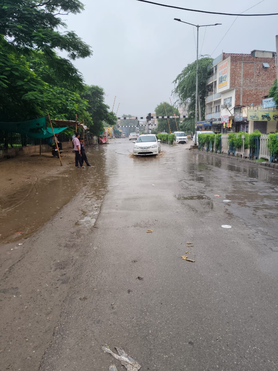 रेलवे फाटक के पास जलभराव, जहां से गुजरते वाहन, पास में खड़े बच्चे।