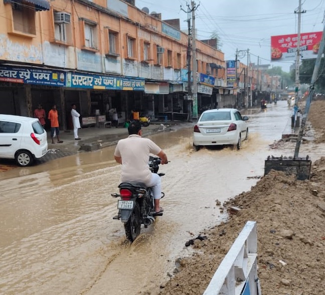 जनता भवन रोड पर जलभराव से साइड में खड़े लोग, पानी से गुजरते वाहन चालक।