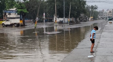 अनाज मंडी में जलभराव, चप्पल पहनकर रास्ता पार खड़ा बच्चा।