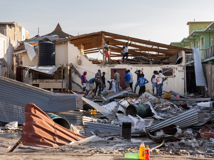 तस्वीर तूफान मेलिसा की वजह से तबाह हुए एक घर की है। लोग मलबे में से खाने-पीने की चीजें और जूस की बोतलें निकाल रहे हैं।
