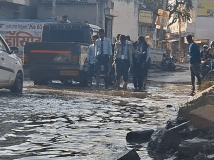 जूतों को हाथ में लेकर गंदे पानी के बीच से स्कूली बच्चे निकल रहे हैं।