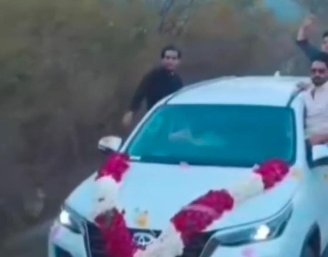 सड़क पर दौड़ती नई फॉर्च्यूनर कार की खिड़की पर लटककर स्टंट करते युवक।