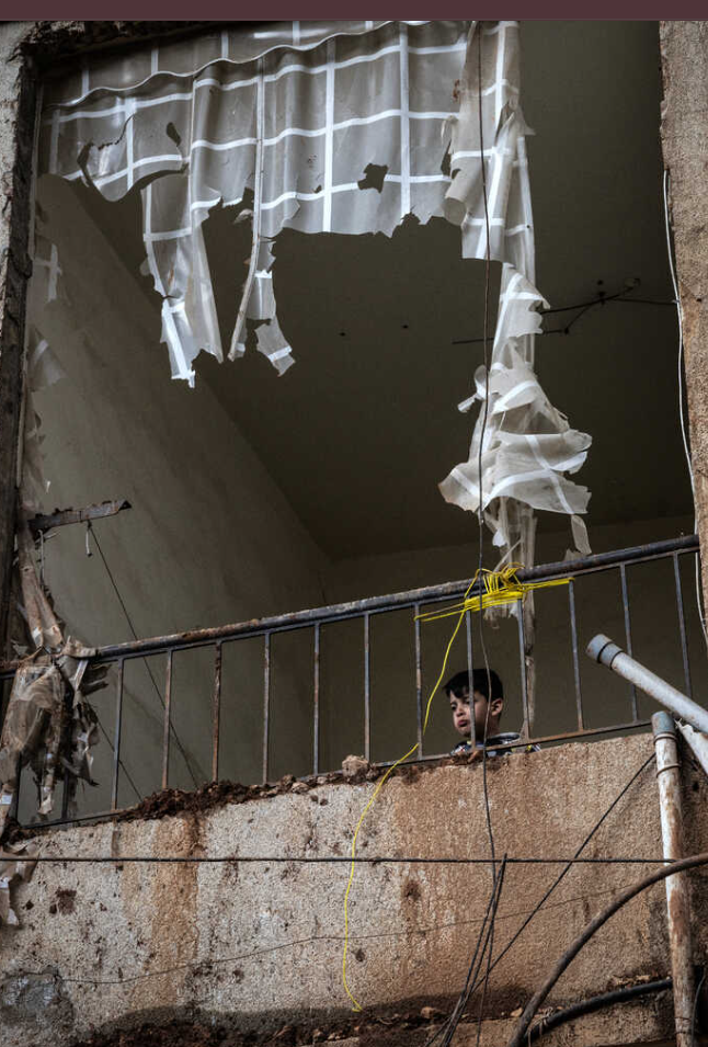 इजराइल के हमले के बाद एक बच्चा टूटी हुई इमारत की बालकनी से झांक रहा है।