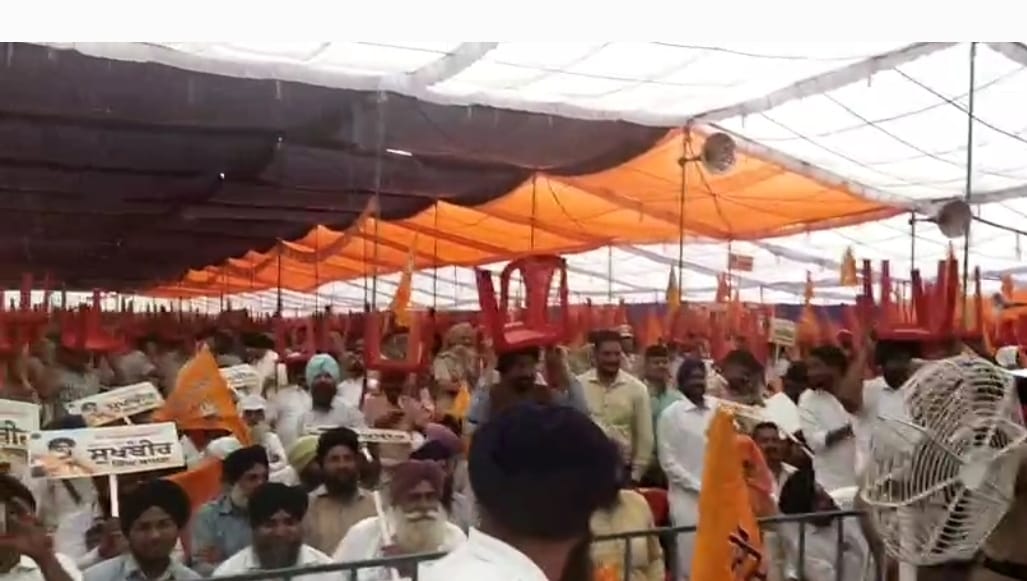 रैली में बारिश आने पर सिर पर कुर्सियां लेकर खड़े लोग।