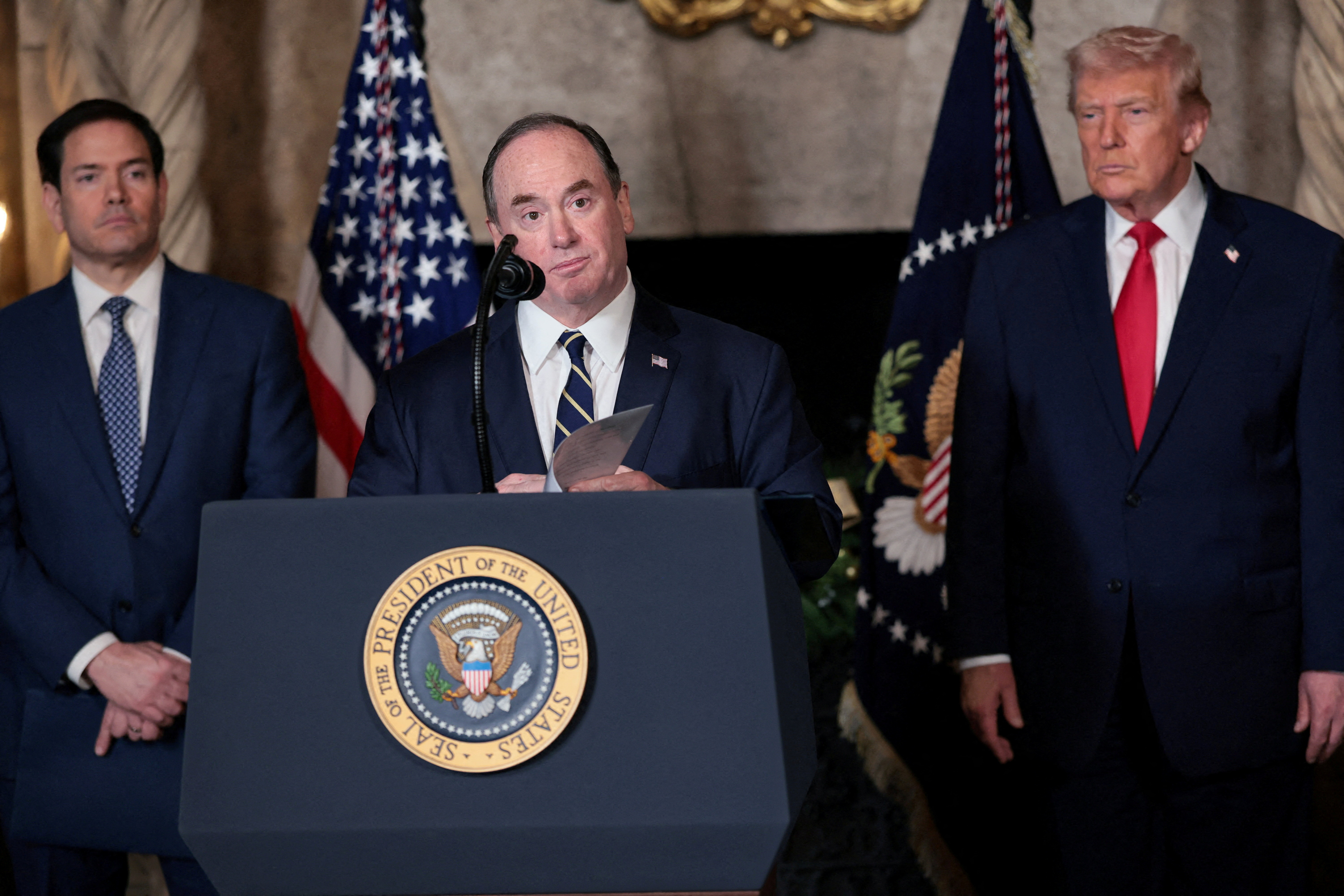 अमेरिका के नेवी सेक्रेटरी जॉन फेलन। उनके पीछे विदेश मंत्री मार्क रूबियो और राष्ट्रपति डोनाल्ड ट्रम्प खड़े हैं।