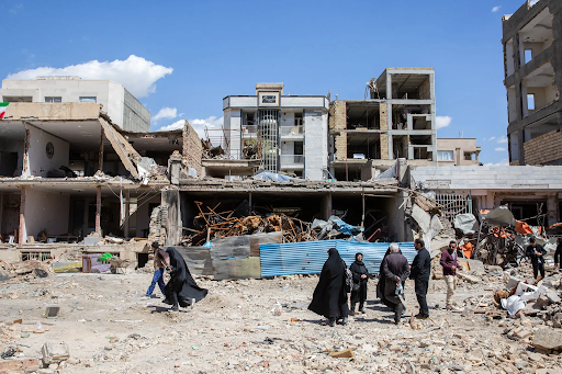 ईरान में क्षतिग्रस्त इमारतों के सामने खड़े लोग। (फाइल फोटो)