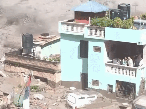 बाढ़ और मलबे के बीच इमारत की बालकनी में तीन लोग खड़े नजर आ रहे हैं।