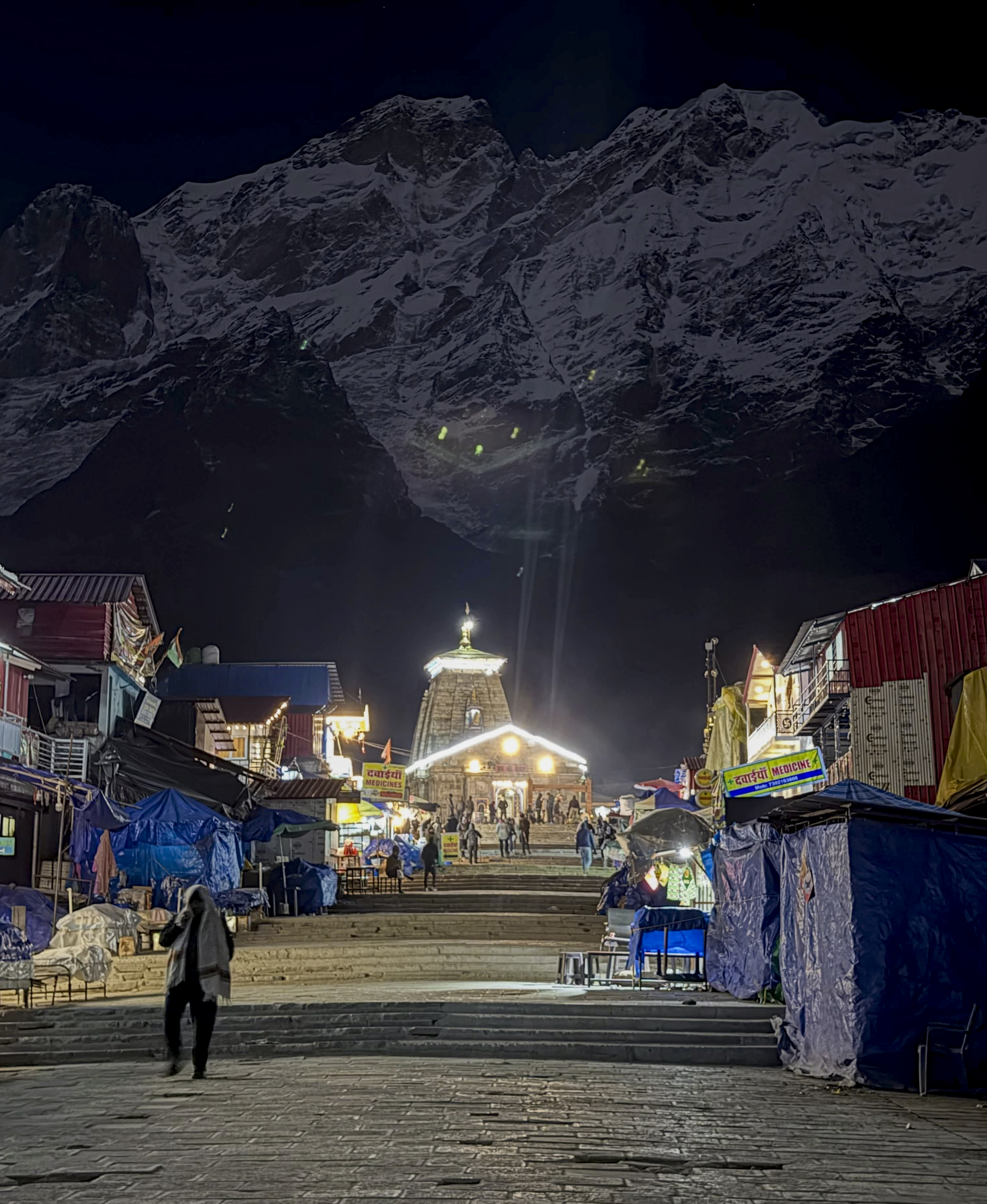 बर्फ से ढके पहाड़ों के बीच रात में रोशनी से जगमगाता केदारनाथ मंदिर।